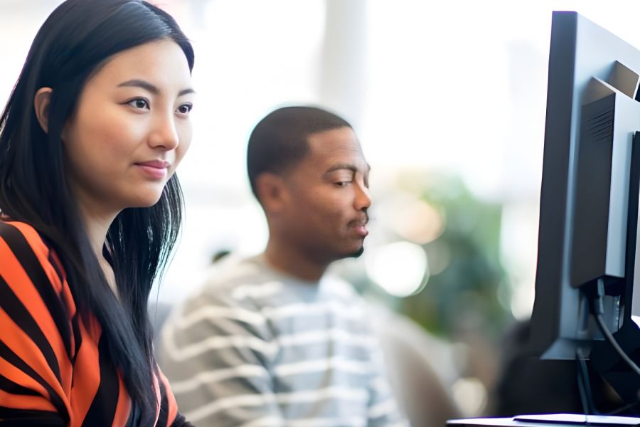 Two people working at desktop computers in a bright, modern office or classroom setting.