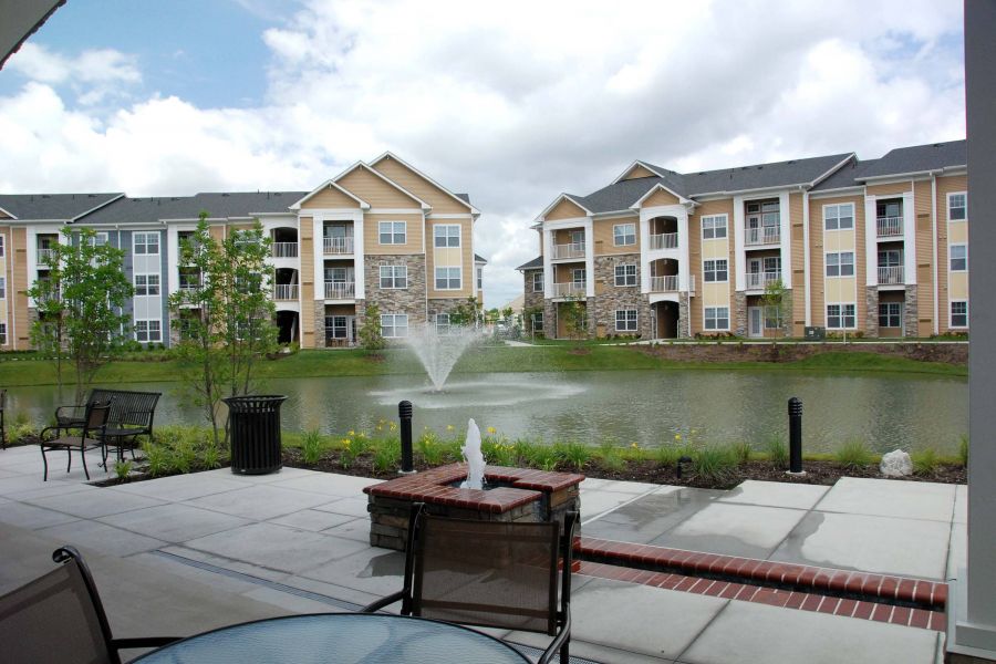 View of apartment buildings and a pond with a fountain, seen from a patio with outdoor seating.