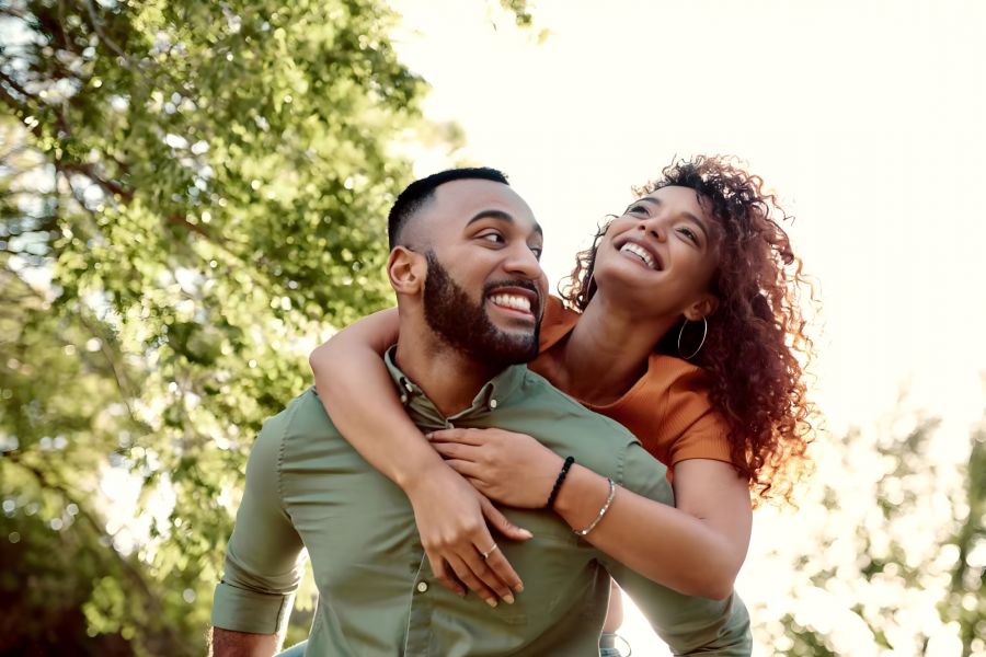 Smiling woman on man's back outdoors, both laughing, surrounded by green trees and sunlight.