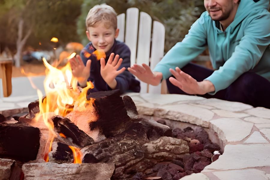 A man and a boy warm their hands by a fire pit outdoors, surrounded by trees and seating.