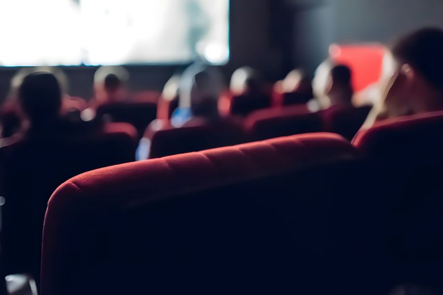 People sit in red seats watching a movie in a dimly lit theater, focus on the back of a chair.