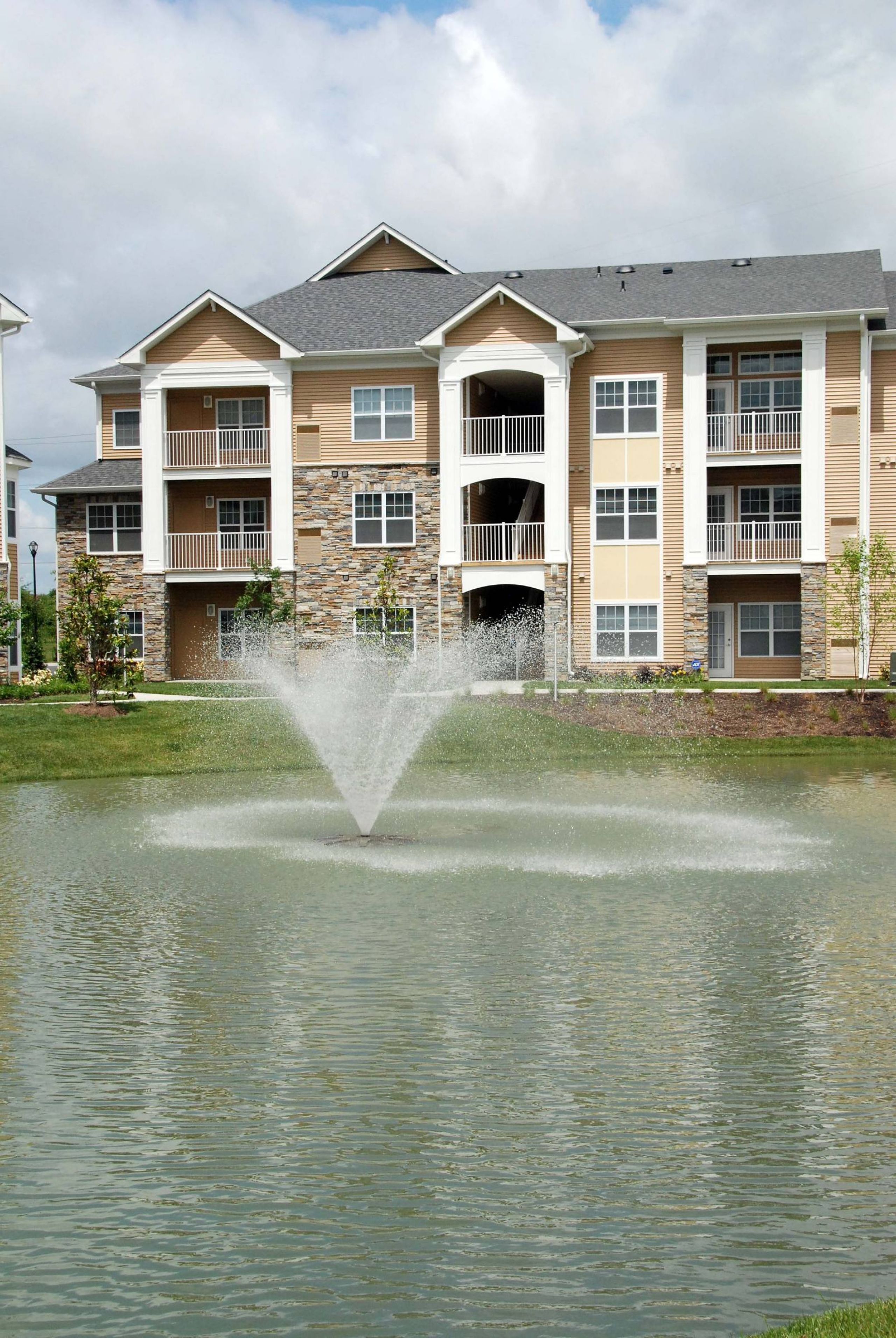 Three-story apartment building with balconies, behind a pond featuring a central water fountain.