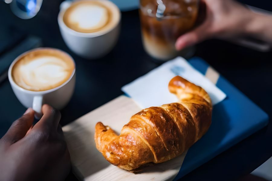 A croissant on a wooden board, two lattes, and iced coffee on a table with people’s hands visible.