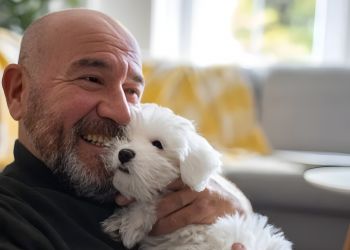 Smiling man cuddling a fluffy white puppy on a sofa in a cozy living room.