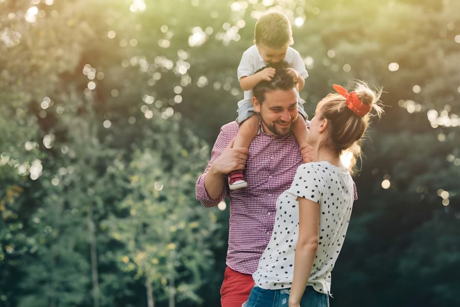 Smiling family with a child on a man's shoulders, outdoors in a sunny, green park.