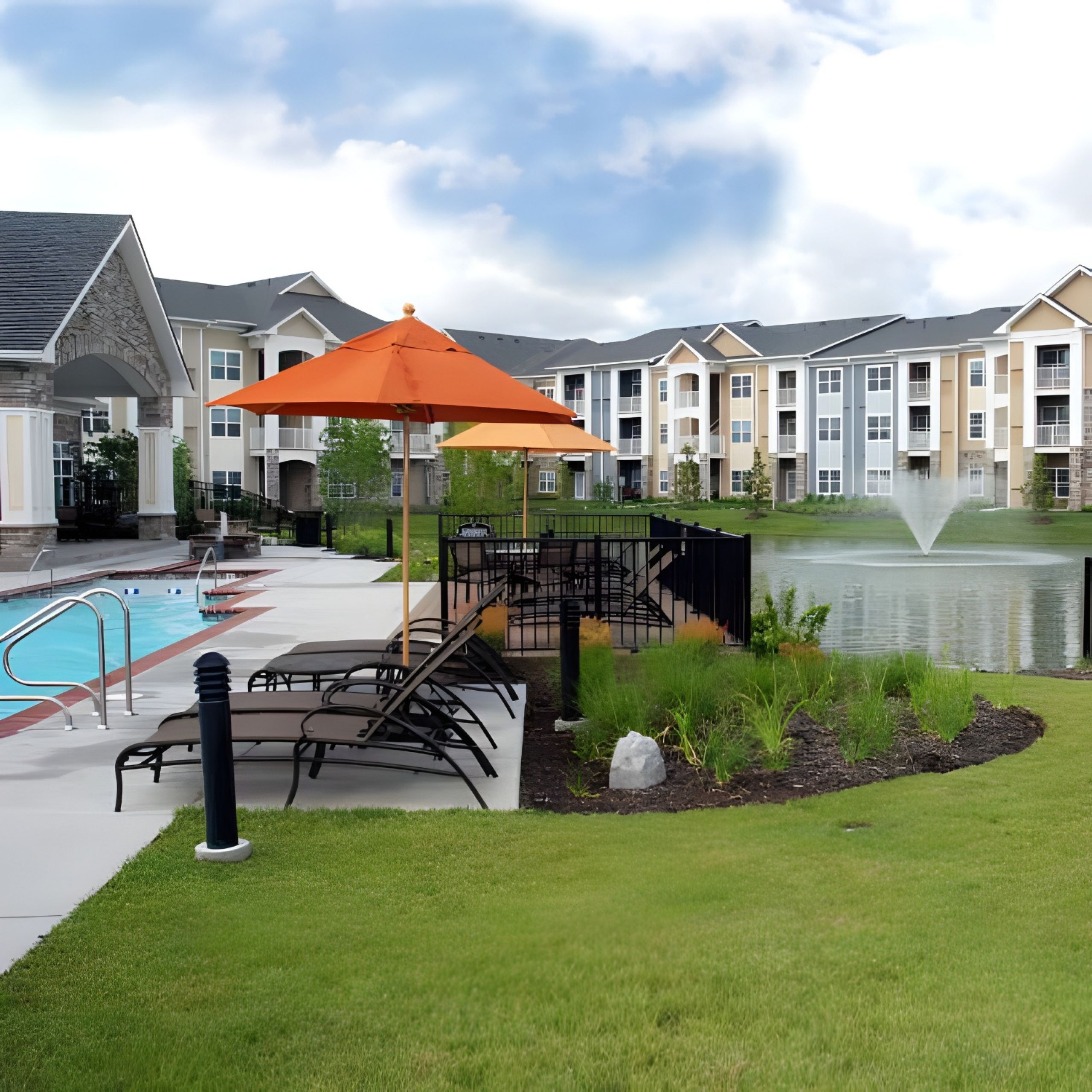 Apartment poolside with lounge chairs, orange umbrella, and a pond with a fountain in the background.