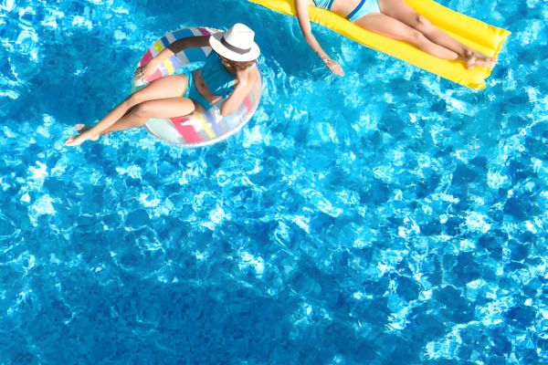 Two women relaxing in a pool, one on a yellow float, the other on a swim ring, both wearing swimsuits and hats.