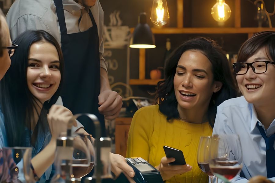 Four friends at a restaurant table smile as a server processes a payment; wine glasses and food on the table.