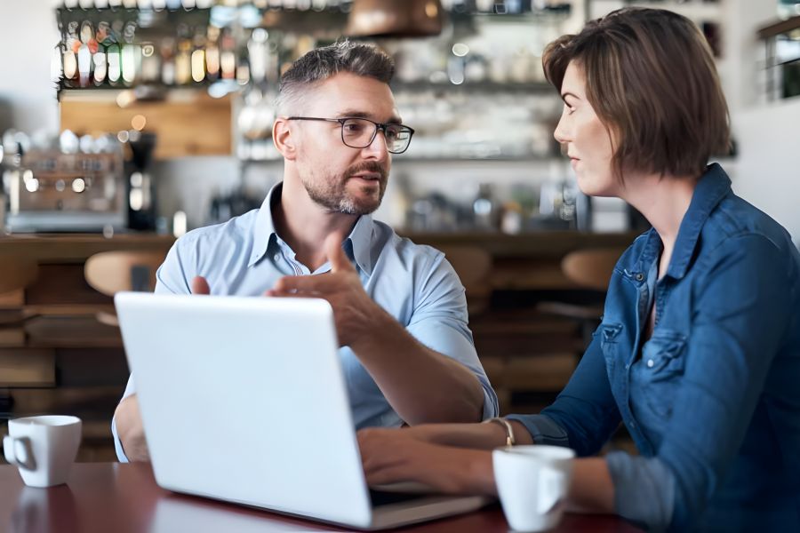 Two people sitting at a café table with a laptop, talking seriously, each with a cup of coffee.