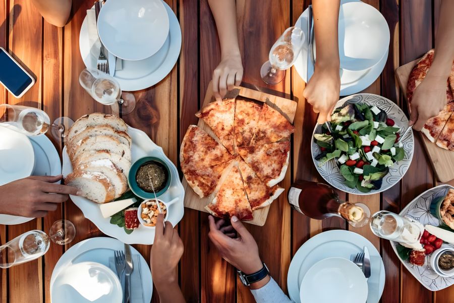 A group shares pizza, bread, salad, and drinks at a wooden table set for a meal.