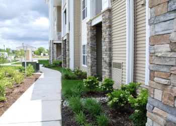 Sidewalk and landscaped greenery along the exterior of a modern apartment building with stone and beige siding.